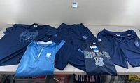 Lot of navy and light blue Rhode Island collegiate basketball apparel on table. Includes shirts, pullover, pants, and shorts, showing branding, logos, and graphics.