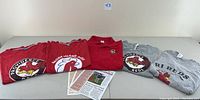 Six Rhode Island Reds and Providence Reds apparel pieces displayed flat on table, including five T-shirts and one polo shirt, in red and gray colors with rooster logos and text. Three large rooster logos stating Providence Reds or Rhode Island Reds are visible on shirts. Two informational cards placed under some shirts.
