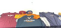 Three shirts and two ball caps arranged in a row against white background, showing logos and colors for Virginia Cavaliers with sizes and colors visible.