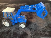 Single blue Ford toy tractor with articulated front loader with grapple seen from side angle on black background.