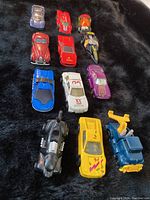 Top view of 12 assorted Hot Wheels toy cars on black background showing variety of colors and styles including red, white, purple, yellow, blue, black, and specialty vehicles