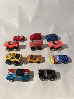 Top-down image showing 11 small toy vehicles including monster trucks and sports cars arranged in rows on a white background.