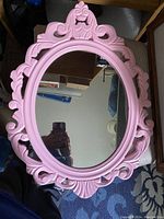 Photo of the pink wooden oval mirror with carved decorative frame placed on a surface reflecting surroundings.