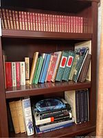 Bookshelves showing rows of multiple books including a full or partial red leather-bound series with gold detailing, combined with other hard and softcover books of various sizes and colors.