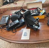 Kodak Six-16 folding camera with lens and user manual, Kodak Instamatic X-15F camera, camera bags and film accessories on table.