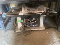 Front view of the Delta ShopMaster 10 inch bench saw with power cord coiled in front, placed on garage floor, some dust and rodent evidence visible.