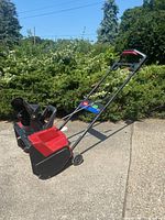 Electric snowblower shown from an angled side view outdoors on a pavement, showcasing the handlebar, chute, and auger section.