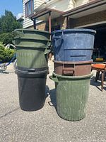 Seven plastic garbage bins stacked outside on driveway in front of garage, including black, green, blue, and brown colors.