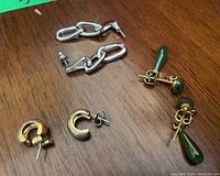 Three pairs of earrings on wooden surface: gold chunky hoops, silver chain links, and gold with green drops.