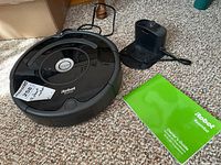 Photo showing front angle of black circular Roomba vacuum robot with CLEAN button on top, docking station with attached power cord, and green owner's guide manual placed on carpet.