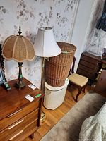 Two lamps positioned beside a wooden dresser. The floor lamp features a white fabric bell-shaped shade and brass-colored base. The shorter table lamp has a dark metal base with a woven wicker-style shade.