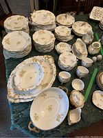 Photo showing the full set of dishes and serving pieces arranged on a table, clearly displaying floral patterns, gold trim, and some chips.