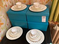 Four place settings of Minton China stacked on blue boxes, including teacups, saucers, appetizer plates, side plates, and dinner plates with gold and silver decorative trim.