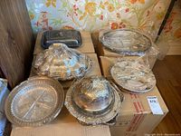 Overview of entire lot showing silver plated covered trays, serving platters wrapped in plastic, and a clear glass serving plate on cardboard boxes.