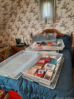 Four clear plastic bins with lids placed on a bed, two of them open showing assorted boxed cards and household items inside.