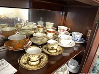 Shelf with eight vintage teacups and saucers of various brands and designs including Noritake demitasse pair, floral and gold trim styles.