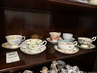 Photo showing 7 teacups with matching saucers displayed on a shelf with floral and ornate patterns