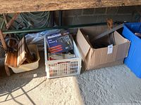 Wide view of assortment under workbench showing MasterCraft drill set in box, plastic crates, and cardboard box with more tools.