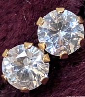 Close-up photo of two round clear cubic zirconia stone stud earrings in 14K yellow gold prong settings on a dark fabric background.