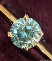 Close-up photo showing the round, brilliant-cut moissanite gemstone held by four prongs on a thin 10K gold band, highlighting the gemstone's clarity and sparkle.