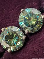 Close-up front view of certified 14K gold moissanite earrings showcasing round brilliant stones in prong settings on purple fabric.