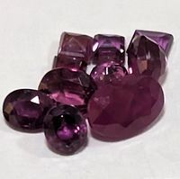 Top view of eight faceted gemstones including one large deep pink-red oval ruby, and seven smaller purple amethysts in various cuts.