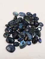 Photo of approximately 38 loose sapphire gemstones in various cuts and sizes with dark blue to dark indigo hues.