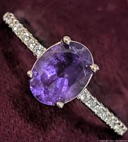 Close-up front view of sterling silver ring with oval amethyst center and CZ encrusted band on a burgundy fabric background.