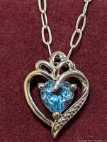 Front view of the sterling silver heart-shaped pendant with a blue topaz gemstone and clear accents on a 16-inch silver chain against a red background.