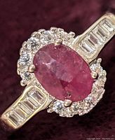 Close-up of the oval ruby center stone surrounded by round cubic zirconia stones and baguette-cut cubic zirconia on the band, showing the ring details and setting.
