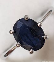 Close-up image showing oval faceted dark blue sapphire gem set in sterling silver band with six prongs.