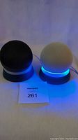 Pair of black and white Amazon Echo Dot smart speakers powered on with blue LED rings, on white tablecloth surface.