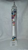 Full view of a 17-inch tall clear glass Galileo thermometer, showing multiple colorful floating glass bubbles with attached metal tags inside a liquid-filled glass tube.