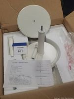 Unassembled white wood round tiers, central columns, and acorn finial topper placed in a box with white foam holders, along with assembly instruction sheet.