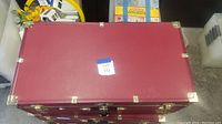 Top and front view of red wood steamer trunk with brass trims and corners, with blue auction lot label.