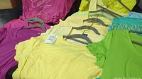 Variety of short sleeve tops in yellow, pink, green, purple, and black, displayed with hanger included.