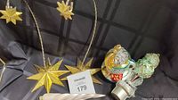 Two goldtone star-shaped ornament displays shown with hanging decorative ornaments in front. Star displays approximately 10.5 inches tall.