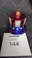 Front view of Radko Liberty Lid glass ornament showing red and white striped top hat with blue brim and white stars, sitting on MaxSold bid card.