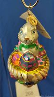 Front angle showing white bunny with blue eyes, green glitter bow tie, colorful glittered Easter eggs, hanging loop with Radko medallion and original tag.