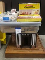 View of unfinished Pueblo style doll house sitting on wooden base with clear plastic tubs containing miniature supplies on top and two unopened yellow Realistic Miniatures furniture kits stacked horizontally.