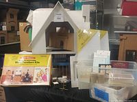 View of unfinished wood doll house model with pitched roof and interior construction. Visible boxed wood furniture kits in front, small tubs, and miniature ceiling panels.