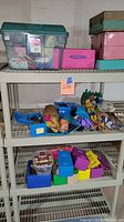 View of three metal shelves with translucent green container, pink and green decorative storage boxes on top shelf. Middle shelf has multiple Barbie dolls and accessories laid out. Bottom shelf has small colorful boxes including blue, yellow, and purple.
