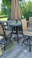 Full view of round high-top patio table with umbrella and surrounding sling chairs on outdoor wooden deck.