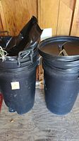 Two black 50-gallon plastic trash cans side by side inside a wooden shed; one contains a black trash bag and miscellaneous items, the other with lid inside the can.