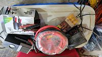 Top view of lot showing car charger box, battery charger, jumper cables coiled in red plastic wrapping, and other items on a white table.
