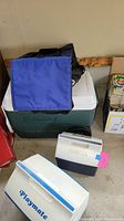Photo showing a large green Coleman cooler with white lid, blue insulated bag on top, and two Igloo Playmate coolers in front.