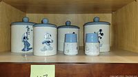 Full set of five Disney ceramic storage containers displayed on a shelf, showing three larger containers with Disney character illustrations and two smaller containers with blue tops and kitchen motif designs.