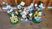 Photo of seven Disney character yard statues on wooden floor showing wear and fading, arranged closely with a wood paneled wall background.