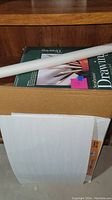 Photo showing a Strathmore Drawing Paper pad, rolled white paper tube, and drawing board in the background.