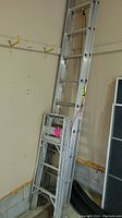 Aluminum extension ladder shown folded and placed in garage corner. Ladder has red tipped feet and visible labels.